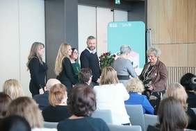 Pokaż zdjęcie: Zdjęcie z tyłu sali zza krzeseł, widać kolejkę osób stojących do Pani Doroty Dutkiewicz, żeby złożyć życzenia