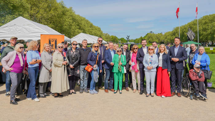 Pokaż zdjęcie: Grupa kilkudziesięciu osób pozuje wspólnie do zdjęcia na świeżym powietrzu, na tle namiotów rozstawionych w parku podczas festynu z okazji Dnia Godności Osób z Niepełnosprawnością Intelektualną. Wśród uczestników znajdują się przedstawiciele organizatorów, goście, opiekunowie i mieszkańcy Szczecina. W tle widoczne są polskie flagi oraz rzeźba Jana Pawła II.