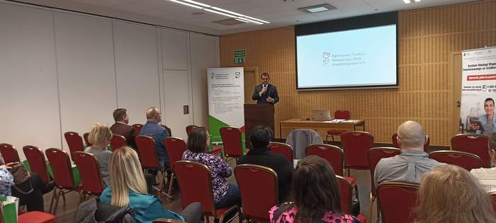 Pokaż zdjęcie: Dyrektor Oddziału Lubuskiego PFRON Andrzej Gonia podczas wystąpienia w sali, na tle logo PFRON i rollup CIDON, stoi przy mównicy, na pierwszym planie goście