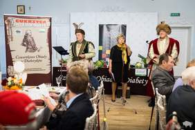 Pokaż zdjęcie: Na zdjęciu dwie osoby w tradycyjnych strojach szlacheckich stoją obok kobiety przemawiającej do mikrofonu. W tle widoczne są banery promujące program "Cukrzyca a Zdrowie" oraz stół z warzywami. Na pierwszym planie znajdują się uczestnicy siedzący przy stołach.