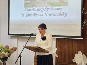 Pokaż zdjęcie: Kobieta w eleganckim, białym żakiecie przemawia przez mikrofon, stojąc naprzeciw mównicy.