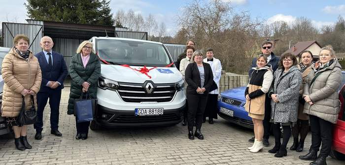 Pokaż zdjęcie: Obok białego mikrobusu stojącego na kostce brukowej po prawej i lewej stronie stoją ludzie. Ubrani są w kurtki. Za nimi stoją auta osobowe. Na masce przedniej asuta czerwona kokarda i niebieska naklejka z poktogramem osoby na wózku.