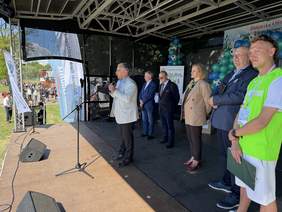 Pokaż zdjęcie: Na scenie plenerowej mężczyzna przemawia do uczestników wydarzenia przy mikrofonie. Obok stoją organizatorzy i goście. W tle widoczne są balony, banery oraz publiczność zgromadzona przed sceną.