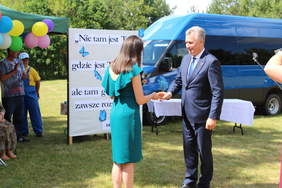 Pokaż zdjęcie: Mężczyzna w garniuturze przekazuje klucze do busa kobiecie w zielonej sukience, w tle granatowy bus z czerwoną kokardą