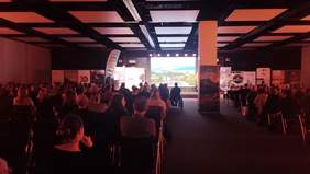 Pokaż zdjęcie: Grupa kilkudziesięciu osób siedzi w sali konferencyjnej. Z przodu sali znajduje się duży ekran, na którym wyświetlany jest film podróżniczy. Po obu stronach ekranu ustawione są banery partnerów 12. Festiwalu Podróżniczego Śladami Marzeń.