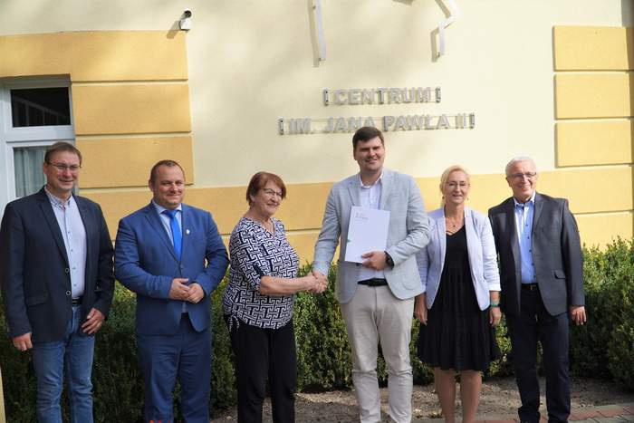 Pokaż zdjęcie: Zdjęcie pozowane przed budynkiem. Przewodnicząca PSONI koło w Nidzicy Ewa Gałka ściska dłoń dyrektora PFRON Mateusza Szkaradzińskiego