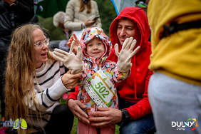 Pokaż zdjęcie: Zbliżenie na małe dziecko w kolorowej kurtce i kapturze, siedzące na kolanach mężczyzny w czerwonej bluzie. Kobieta w okularach i swetrze z paskami pomaga dziecku założyć beżowe rękawiczki. Dziecko uśmiecha się, ma przypięty numer startowy 502. Wszyscy są na zewnątrz.