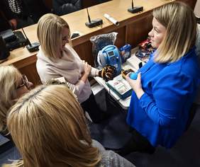 Pokaż zdjęcie: Cztery kobiety stoją pochylone nad małym stolikiem, oglądając sprzęt rehabilitacyjny i pomoce. Jedna z nich w niebieskiej marynarce trzyma niebieską kulkę z wypustkami. Na stoliku widać niebieskie urządzenie medyczne oraz pomarańczowo-czarną rękawicę do ćwiczeń.