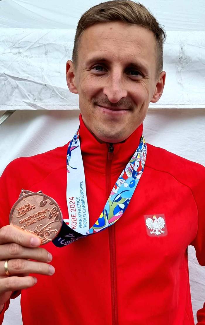 Pokaż zdjęcie: Maciej Lepiato prezentuje brązowy medal, który trzyma w ręce, jest uśmiechnięty, ubrany w czerwony dres z orzełkiem na piersi