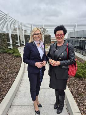 Pokaż zdjęcie: Dwie kobiety stoją na ścieżce na zewnątrz budynku, uśmiechając się do aparatu. Jedna z nich ma blond włosy i nosi granatowy garnitur z białą koszulą oraz apaszką, druga – czarne włosy, okulary w czerwonych oprawkach oraz czarny skórzany strój z czerwoną torbą. W tle widać nowoczesną architekturę z białymi, metalowymi elementami i roślinnością.