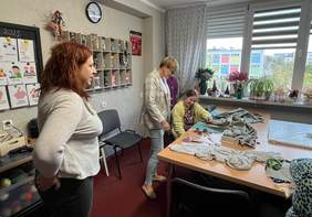 Pokaż zdjęcie: Trzy kobiety w jasnym pomieszczeniu, dwie z nich pochylone nad stołem z materiałem i przyborami do szycia. W tle okno i tablica na ścianie z rysunkami.