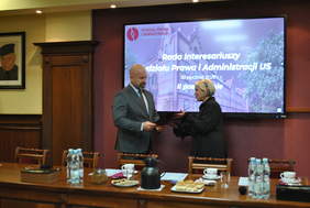 Pokaż zdjęcie: Mężczyzna w garniturze wręcza dokument kobiecie stojącej obok niego przed stołem prezydialnym. Za nimi ekran z napisem „Rada Interesariuszy Wydziału Prawa i Administracji US, 30 stycznia 2026 r., II posiedzenie”. Na stole widoczne są mikrofony, woda i poczęstunek.