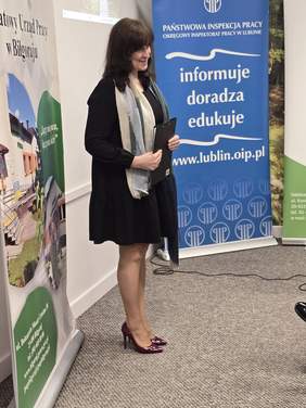 Pokaż zdjęcie: Wysoka, szczupła kobieta, ubrana w czarną sukienkę w kolano podczas prezentacji.