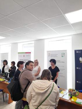 Pokaż zdjęcie: Stoisko informacyjne, na stolikach leżą ulotki, za stolikami banery z logo PFRON, OWIT, NFZ, na pierwszym planie sylwetki osób odwiedzających stoiska.