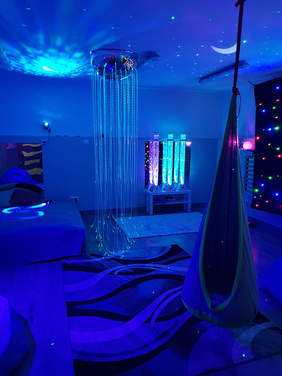 Pokaż zdjęcie: Duży pokój w którym włączone są kolorowe światełka imitujące noc, punkty świetlne, gwiazdozbiór. W lewym rogu stoi rozłożona kanapa przykryta kocem. Po prawej stronie ściana ma ciemnogranatowy kolor, a na niej świecą się kolorowe punkty g