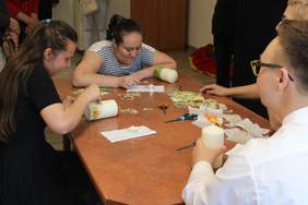 Pokaż zdjęcie: Przy stole siedzą dwie dziewczynki i chłopiec. Każdy ma w ręku świeczkę, którą ozdabiat