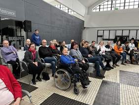 Pokaż zdjęcie: Na jasnoszarych fotelach siedzą ludzie uśmiechają się do aparatu. Na pierwszym planie jest  kobieta na wózku inwalidzkim.