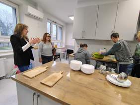 Pokaż zdjęcie: Dwie osoby stojące tyłem przygotowują posiłek, w kuchni na blacie stoją talerze oraz leżą deski.