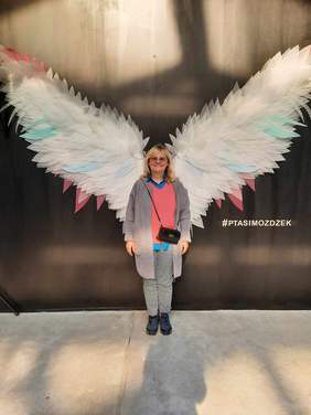 Pokaż zdjęcie: Przed ścianką do robienia zdjęć stoi kobieta, która uśmiecha się szeroko. Na ściance znajdują się ogromne opierzone skrzydła. 