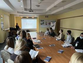 Pokaż zdjęcie: W Sali konferencyjnej przy długim drewnianym stole siedzi kilka osób. Patrzą się w stronę dużego wiszącego białego ekranu na którym wyświetla się logo PFRON. Przy ekranie stoi kobieta w długich blond włosach i ciemnej sukience która mówi do