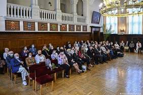 Pokaż zdjęcie: Duża sala, na podłodze lśni parkiet , na ścianach obrazy i herby. Z sufitu zwisa duży złoty żyrandol. Po jednej stronie siedzą na drewnianych krzesłach galowo ubrani ludzie oraz osoba  na wózku inwalidzkim.