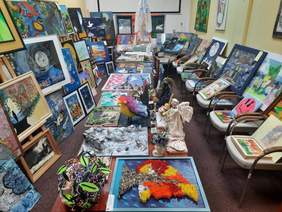 Pokaż zdjęcie: W sali konferencyjnej znajduje się mnóstwo prac plastycznych nadesłanych w ramach konkursu artystycznego "Sztuka Osób Niepełnosprawnych". Obrazy stoją na sztalugach i krzesłach. Na stołach leżą rzeźby. W pomieszczeniu jest bardzo barwnie i kolorowo. 