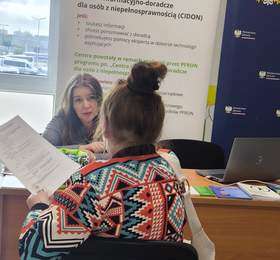 Konsultacja ekspercka PFRON – dwie osoby siedzą przy biurku i rozmawiają z interesantem trzymającym dokument; w tle roll‑up CIDON, laptop oraz materiały informacyjne na blacie.