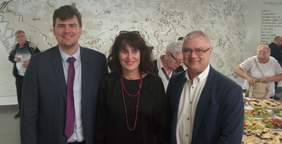 Pokaż zdjęcie: Mężczyzna po lewej stronie to młody uśmiechnięty brunet, po prawej stoi uśmiechnięty pan w marynarce i okularach. Po środku uśmiechnięta kobieta o ciemnych włosach. W tle suto zastawiony stół i uczestnicy, którzy częstują się poczęstunki