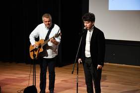 Pokaż zdjęcie: na scenie stoi dwóch mężczyzn, pierwszy od lewej ubrany w jasną koszulę i ciemne spodnie, szpakowaty gra na gitarze, obok stoi młody mężczyzna w marynarce i śpiewa.