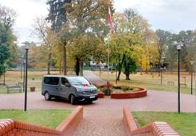 Pokaż zdjęcie: bus w kolorze szarym metalicznym na tle posesji w kształcie parku, są drzewa, jesienna aura, żółte liście, na pierwszym planie także masz z flagą Polski