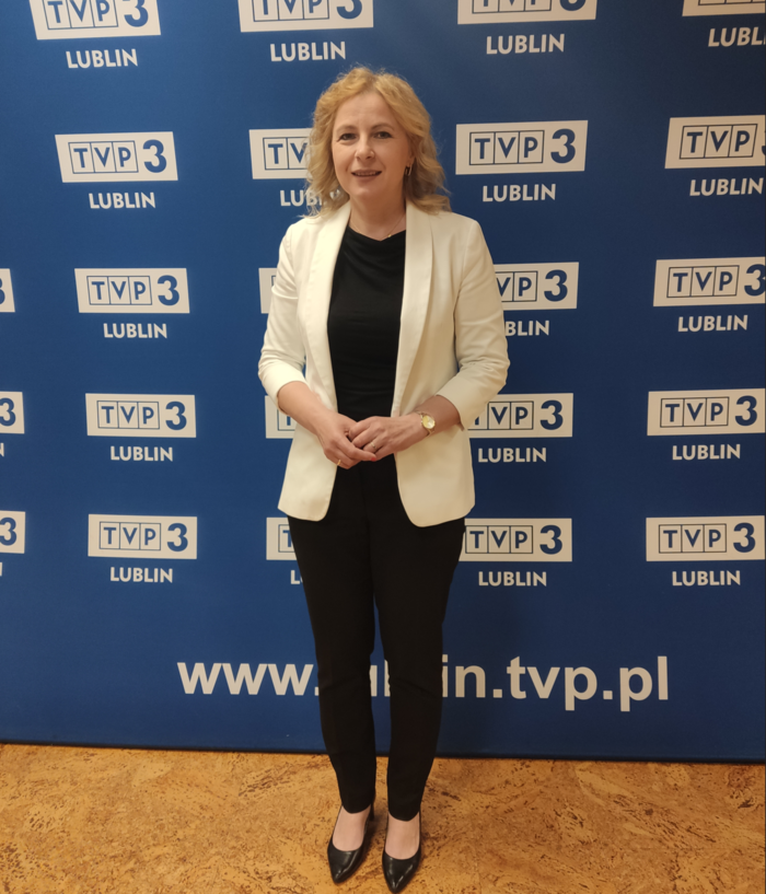 Pokaż zdjęcie: Na tle banneru z logo TVP3 Lublin, pozuje kobieta blondynka ubrana w czarną bluzkę i spodnie oraz w białą marynarkę.