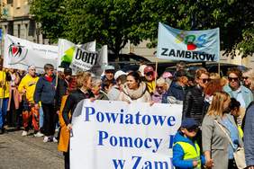 Pokaż zdjęcie: Grupa osób uczestniczy w marszu integracyjnym. Na zdjęciu widać tłum ludzi idących ulicą, niosących transparenty z nazwami instytucji wsparcia społecznego, m.in. „ŚDS Zambrów”, „Dom Pomocy Społecznej w Białymstoku” oraz „Powiatowe Centrum Pomocy Rodzinie w Zambrowie”. Uczestnicy są ubrani w kolorowe kurtki, część nosi odblaskowe kamizelki. Panuje słoneczna pogoda, a marsz odbywa się w otoczeniu miejskiej zabudowy i drzew.