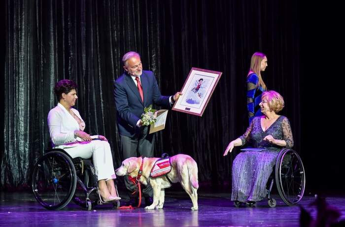Pokaż zdjęcie: Mężczyzna ubrany w garnitur trzyma ramę z dyplomem. Pokazuje ją kobiecie ubranej w jasny garnitur siedzącej na wózku. Obok stoi jasny pies przewodnik. Po prawej stronie mężczyzny kobieta ubrana w suknię wieczorową siedząca na wózku przygląda 