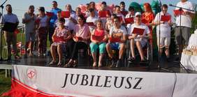 Pokaż zdjęcie: scena w plenerze na której siedzą na krzesłach osoby, trzymają w ręku czerwone arkusze 