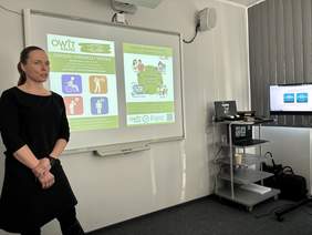 Pokaż zdjęcie: W centralnej części slajdu widać kobietę w czarnym stroju, stojącą przy tablicy projektowej i mówiącą do grupy. W tle jest wyświetlana prezentacja na tablicy. Po prawej stronie sali stoi wózek ze sprzętem i monitor, a przy oknie są zasłony