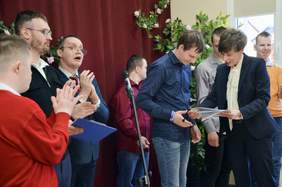 Pokaż zdjęcie: Moment wręczenia dyplomu na scenie. Po prawej kobieta w ciemnej marynarce przekazuje dyplom młodemu mężczyźnie, który w skupieniu patrzy na dokument. Na środku stoi mikrofon, a po lewej grupa osób bije brawo. W tle bordowe zasłony udekorowane kwiatami i liśćmi.