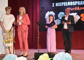 Pokaż zdjęcie: Udekorowana scena, na której stoją cztery osoby. Kobieta przy mikrofonie składa życzenia.