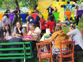 Pokaż zdjęcie: Barwni uczestnicy Miodobrania-Miodogrania w Konarach.  Osoby z niepełnosprawnościami siedzą przy stolikach i wykonują prace plastyczne. w tle widać tańczące osoby. Cześć uczestników jest przebranych za pszczoły.