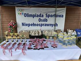 Pokaż zdjęcie: Stół z białym obrysem. Na nim stoją różnego rozmiaru puchary medale i nagrody za nim baner z napisem olimpiada sportowa osób niepełnosprawnych.