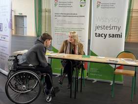 Pokaż zdjęcie: Przy stoisku informacyjnym rozmawiają dwie osoby. Jedna z nich porusza się na wózku. W tle znajdują się materiały informacyjne oraz banery PFRON z hasłem „Wszyscy jesteśmy tacy sami”.