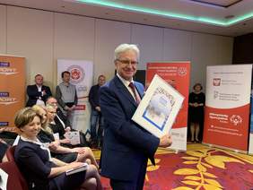 Pokaż zdjęcie: Krzysztof Michałkiewicz, Pełnomocnik Rządu ds. Osób Niepełnosprawnych prezentuje okolicznościową kartkę i znaczek pocztowy wydany przez Pocztę Polską