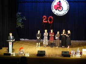 Pokaż zdjęcie: Na scenie znajduje się sześć kobiet oraz jeden mężczyzna, który stoi przy mównicy i przemawia do mikrofonu. Panie biją brawo i uśmiechają się. 