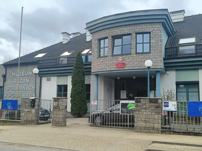Pokaż zdjęcie: Zdjęcie jednopiętrowego budynku w szarej cegle, budynek jest ogrodzony, po lewej stronie na budynku jest duży napis Muzeum Badań Polarnych w Puławach.