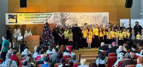 Pokaż zdjęcie: Na scenie stoi liczna grupa dzieci, młodzieży i dorosłych wykonujących kolędy; przed nimi publiczność zgromadzona na sali, obok choinka i baner wydarzenia.