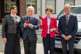 Pokaż zdjęcie: Uroczyste przekazanie kluczy do nowego busa. Przed budynkiem stoją cztery osoby: kobieta w czarnej skórzanej kurtce, mężczyzna w granatowym garniturze trzymający kluczyki z wstążką, kobieta w czerwonej marynarce (również z kluczykami) oraz mężczyzna w ciemnym garniturze i czerwonym krawacie.