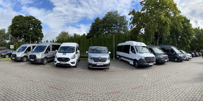 Pokaż zdjęcie: Panoramiczne zdjęcie pokazujące busy z przodu marki Mercedes, Ford, Reno, Opel w kolorach biały, srebrny, brązowy. Busy stoją na kostce brukowej. W tle zielony żywopłot z tui, błękitne niebo i kilka chmurek.