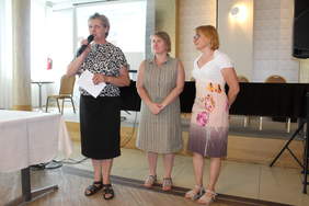 Pokaż zdjęcie: W sali konferencyjnej stoją trzy kobieta, jedna z nich mówi do mikrofonu