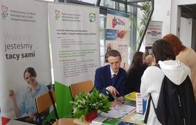 Pokaż zdjęcie: Dwie kobiety oraz mężczyzna udzielający informacji znajdują się przy stoisku Oddziału Wielkopolskiego PFRON. Na stoliku ułożone są materiały informacyjne oraz bukiet konwalii. W tle widoczne są banery: PFRON i CIDON oraz sąsiadujących wystawców.