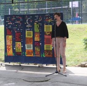 Pokaż zdjęcie: Przy mikrofonie stoi kobieta ubrana w czarną bluzkę i jasnoróżowe spodnie.