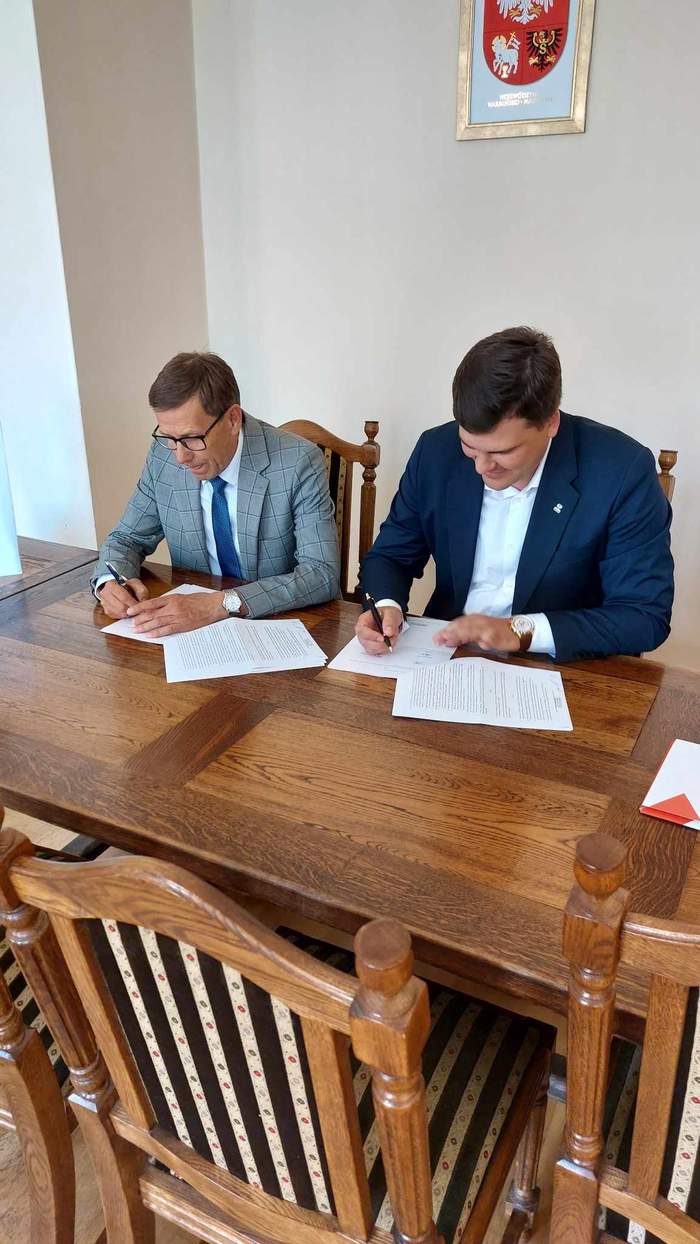 Pokaż zdjęcie: Dwóch mężczyzn w garniturach siedzi przy dużym stole, mają w rękach długopis i podpisują umowy, na ścianie nad nimi herb
